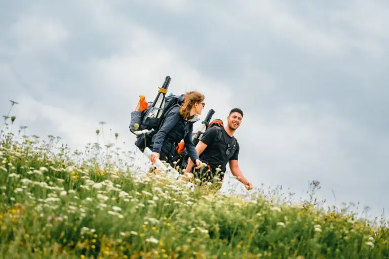 two hikers with large backpacks walking through a wildflower field